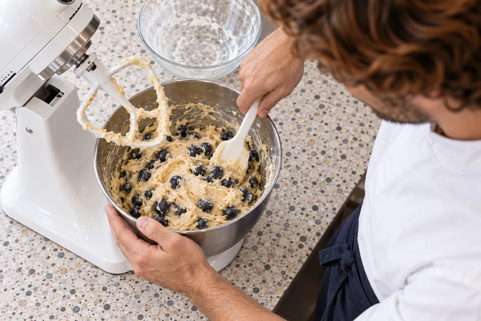 From above, the cook gently folds whole blueberries through thick lemon cookie dough with the mixer stopped.
