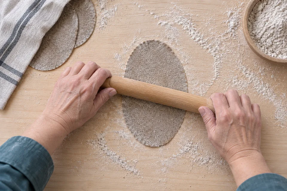 Hands roll a thin oval rye wrapper beside two covered wrappers.