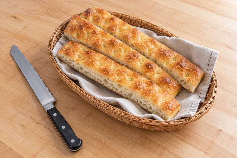 Three thin golden strips of plain focaccia in a shallow linen-lined basket beside a bread knife.