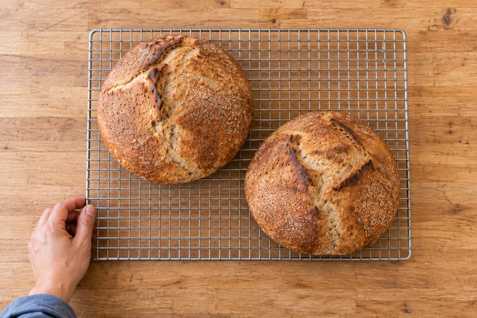 Overhead view of two intact mixed-grain round loaves arranged diagonally on a rack, with one resting hand at its corner.