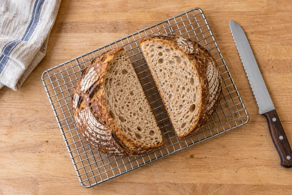 Two matching whole-wheat sourdough halves lie cut-face up on a silver rack, viewed from above beside a bread knife.