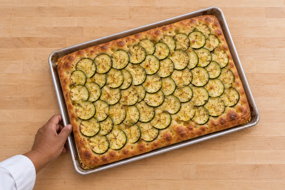 An overhead view of one shallow oblong focaccia covered with overlapping zucchini rounds on its baking sheet.