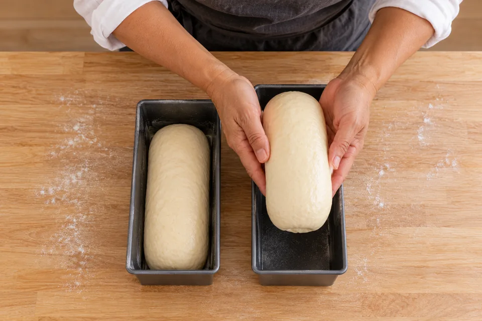 Baker lowers the second smooth dough log into a small loaf tin beside the first filled tin.