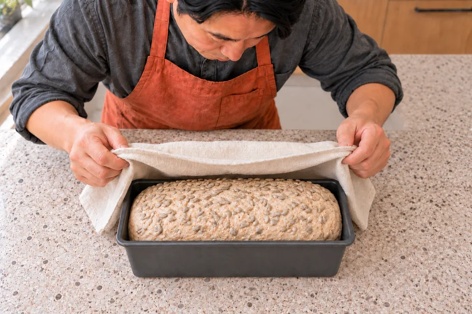A risen raw loaf carries an attached sunflower-kernel coating.