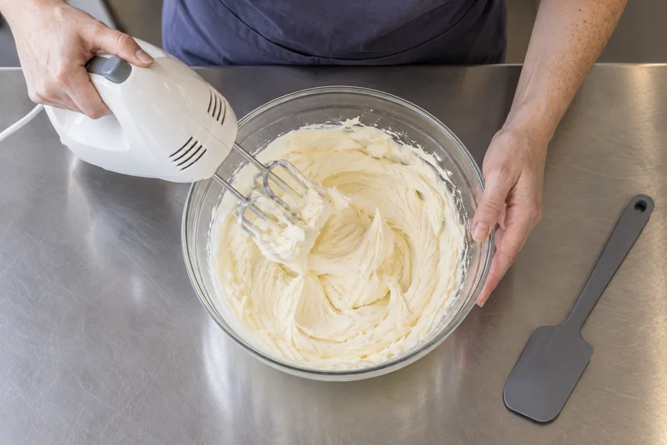 Hands lift mixer beaters from thick cream cheese icing.