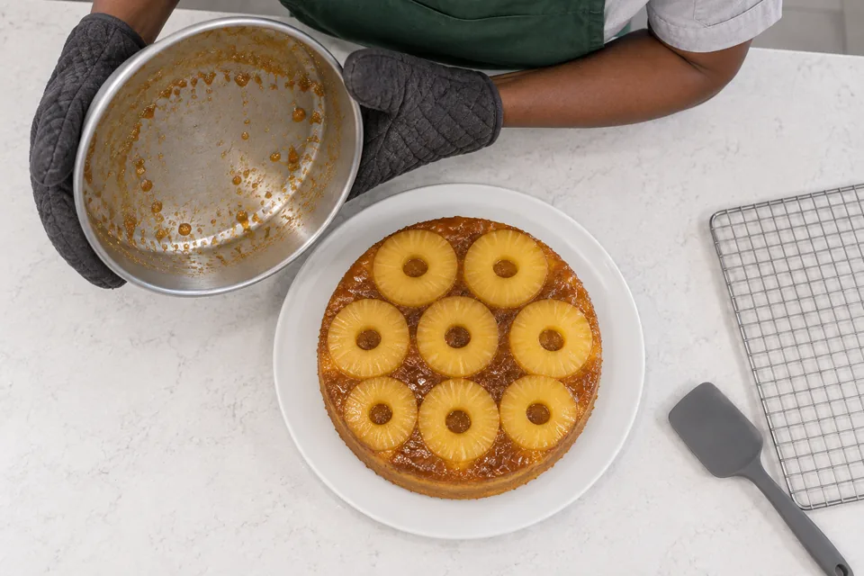 A cook lifts the inverted tin from a pineapple-topped cake on a serving plate.
