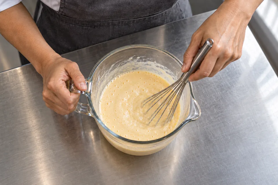Hands whisk smooth pale batter in a clear glass pouring jug.