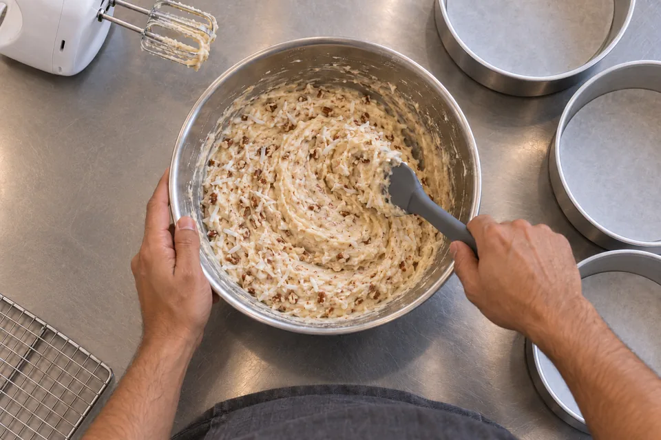 Hands fold coconut and chopped pecans through pale cake batter.