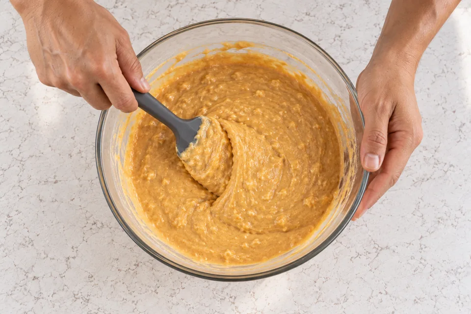 Grey spatula gently folds thick orange pancake batter with small lumps.